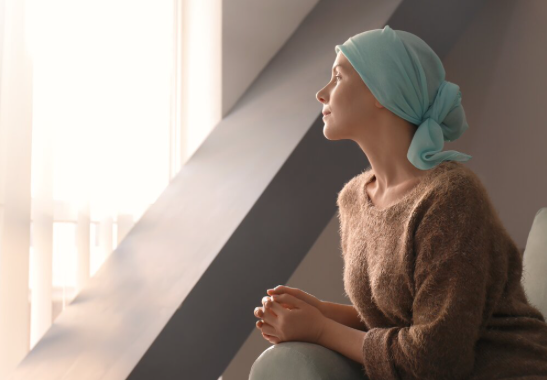 A woman gazes thoughtfully out of a window with her hands clasped together wearing a brown sweater and blue headscarf.