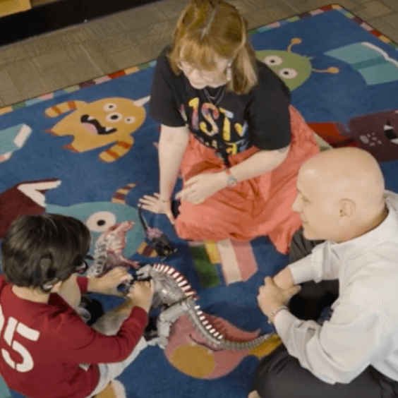 Two adults sit on a floor rug with a deaf student playing.