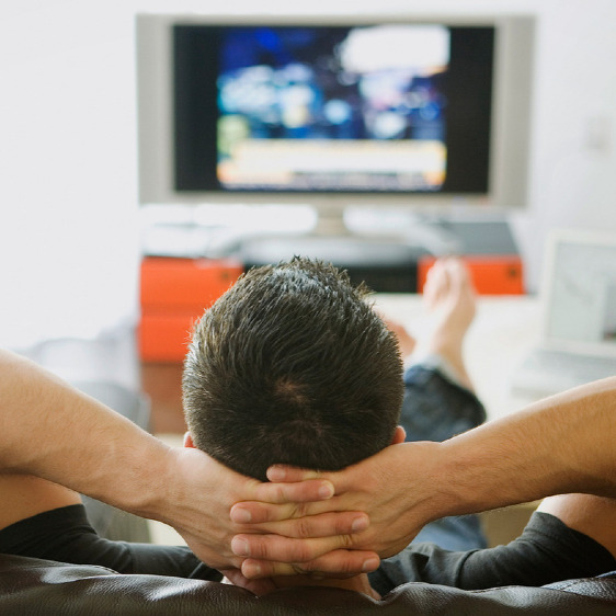 A view from behind of a man with his feet up and hands clasped behind his head watching T.V.